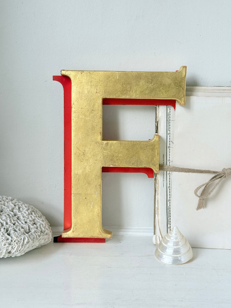 A large decorative letter F with a textured gold finish and red inner panels, displayed on a white surface next to a white box and a small clear glass object.