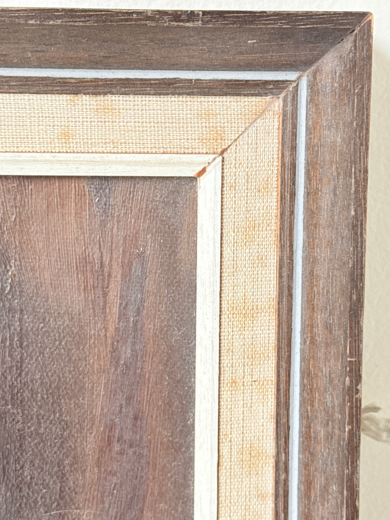Close-up of a framed wall art corner showing layered wood trim in dark brown and medium brown tones with a light beige textured inner border and a thin white edge.