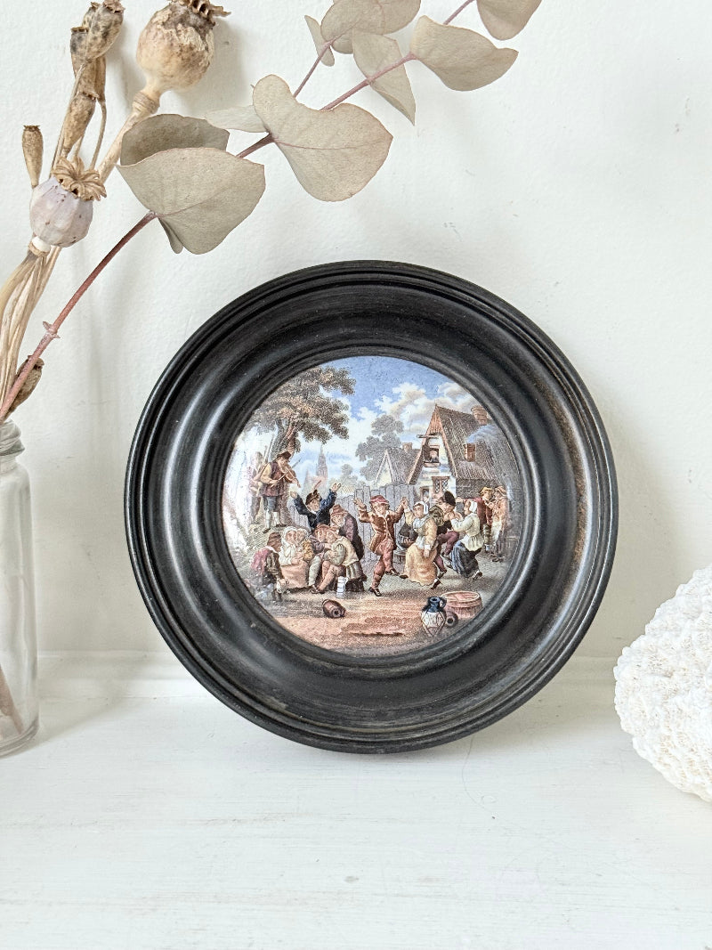 Antique Prattware pot lid in black round frame with rural village scene on white surface.