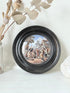 Antique Prattware pot lid in black round frame with rural village scene on white surface.