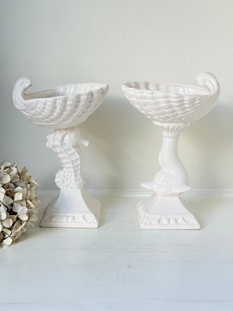 A pair of white ceramic seahorse-shaped pedestal planters with textured bowl-like tops, displayed on a white surface against a light neutral wall.