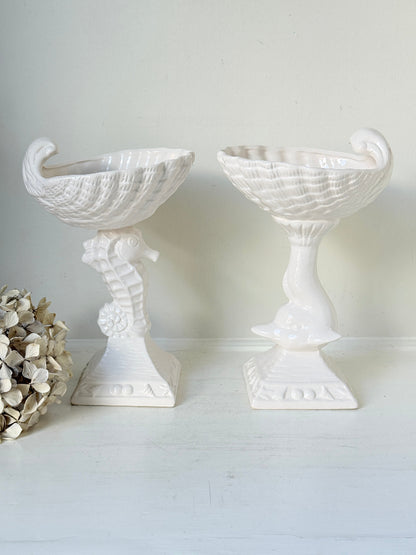 A pair of white ceramic seahorse-shaped pedestal planters with textured bowl-like tops, displayed on a white surface against a light neutral wall.