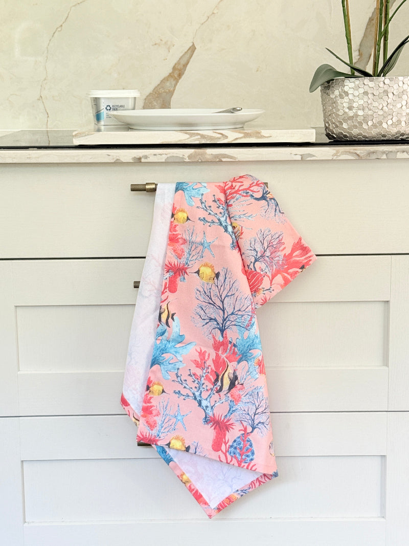 Warm peach coral and tropical fish print cotton tea towel with wooden forks on a marble surface beside cut glass