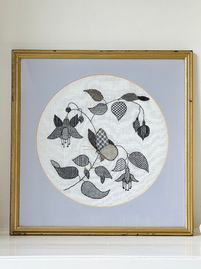 Framed circular hand embroidery of birds and fuchsia flowers in black and grey on white linen, set in gold frame, white surface background.