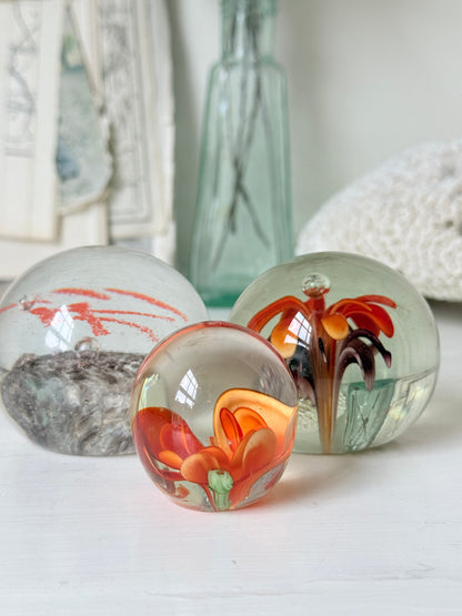 Trio of orange flower glass paperweights with green stems and controlled bubbles, photographed on a white surface