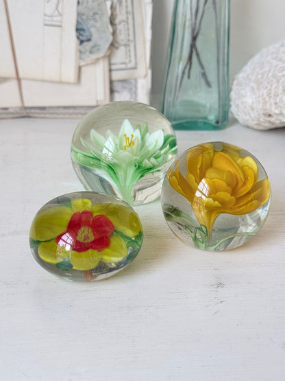 Trio of mixed flower glass paperweights in green, yellow and red, graduated in size, photographed on a white surface