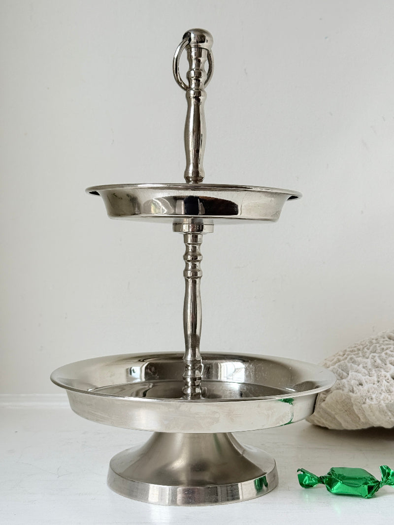 Two tier silver tone cake stand with polished reflective finish styled with green wrapped sweets on a white surface