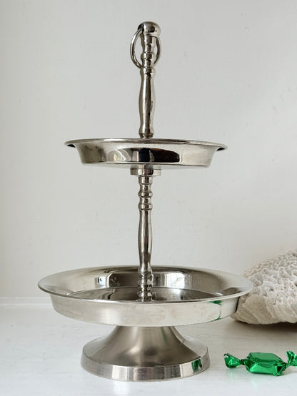 Two tier silver tone cake stand with polished reflective finish styled with green wrapped sweets on a white surface