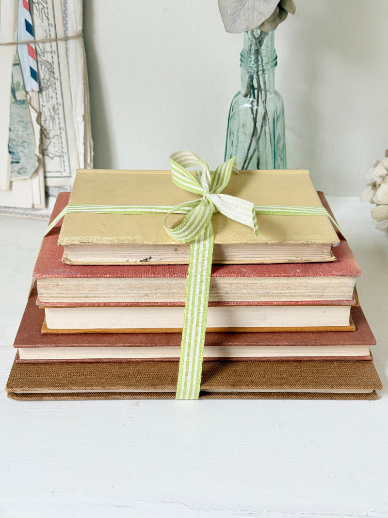 A stack of vintage hardcover books tied with a light green and white striped ribbon, set against a softly blurred background with a glass bottle and framed artwork.