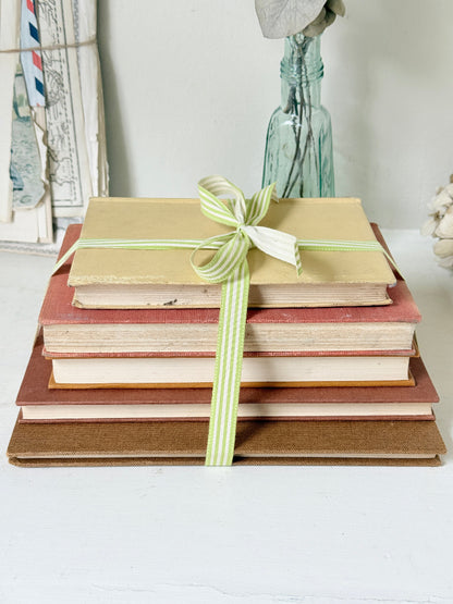 A stack of vintage hardcover books tied with a light green and white striped ribbon, set against a softly blurred background with a glass bottle and framed artwork.