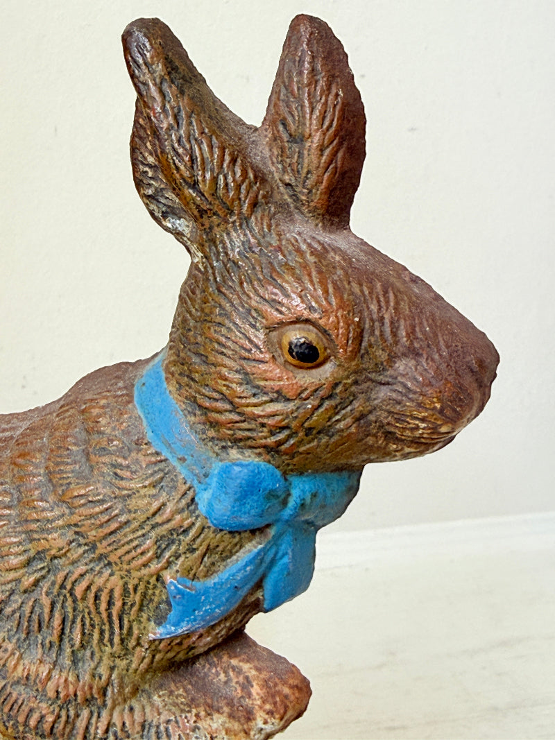 A close-up of a textured brown rabbit figurine wearing a bright blue bow tie, set against a plain off-white background.