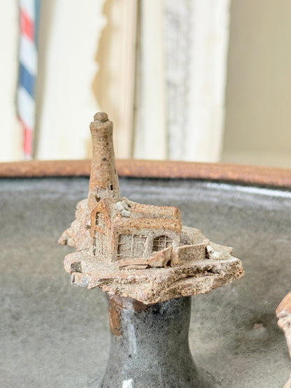 A miniature rustic clay or ceramic building with a tall chimney, displayed on a dark gray ceramic stand.