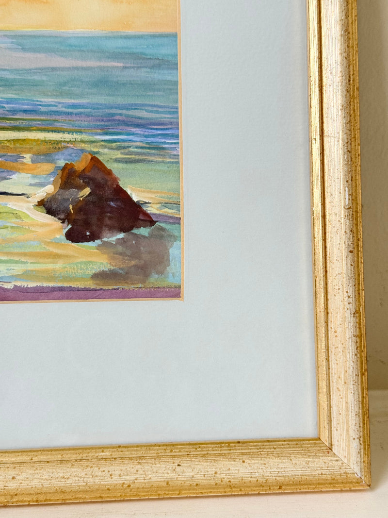 Framed coastal watercolour of sunset over sea with rocks in foreground, in gilt frame with pale mount, photographed on white surface.