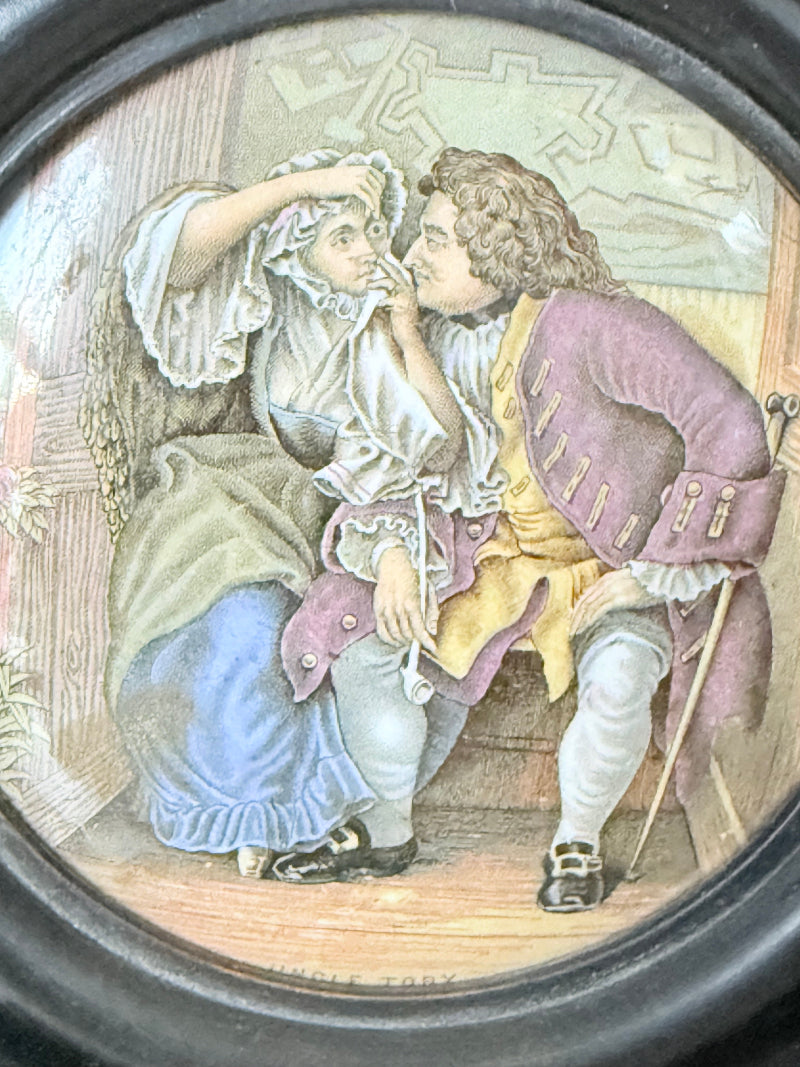 Antique Prattware pot lid in a black round frame with a detailed Victorian scene on a white surface.