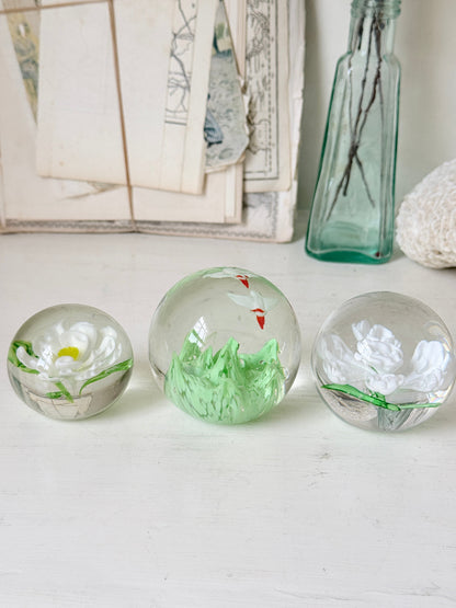 Trio of white flower glass paperweights in graduated sizes with green leaves, photographed on a white surface