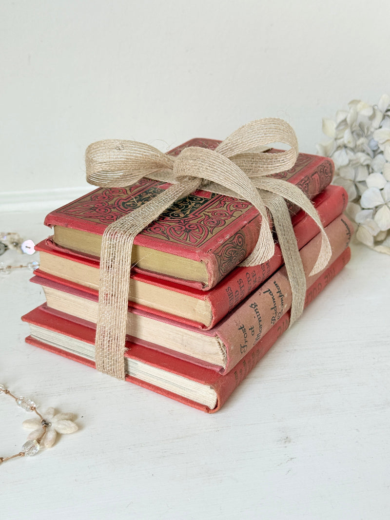 A stack of four vintage-style red books tied with a beige burlap ribbon, photographed on a light tabletop with soft decorative props in the background.