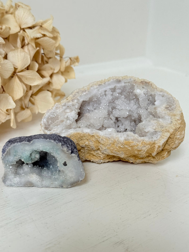 Pair of natural quartz geodes, one white and one lilac-toned, styled on a white surface with dried hydrangea