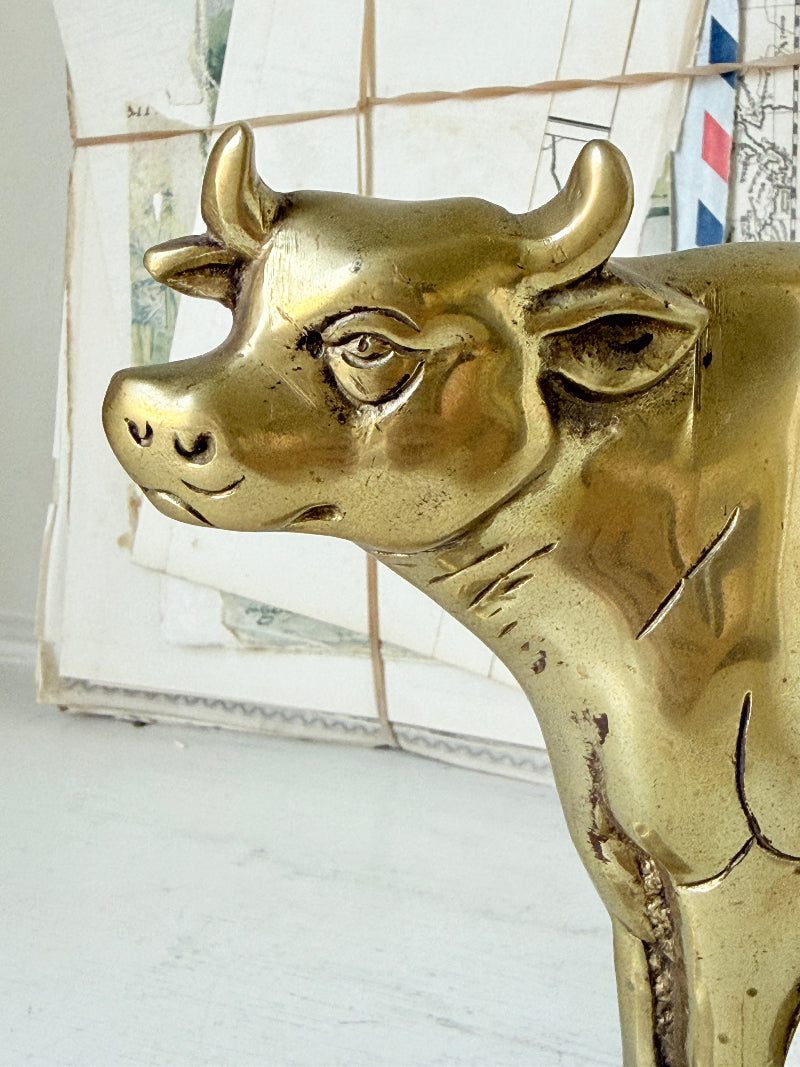 A polished brass cow figurine shown in profile against a white tiled background.