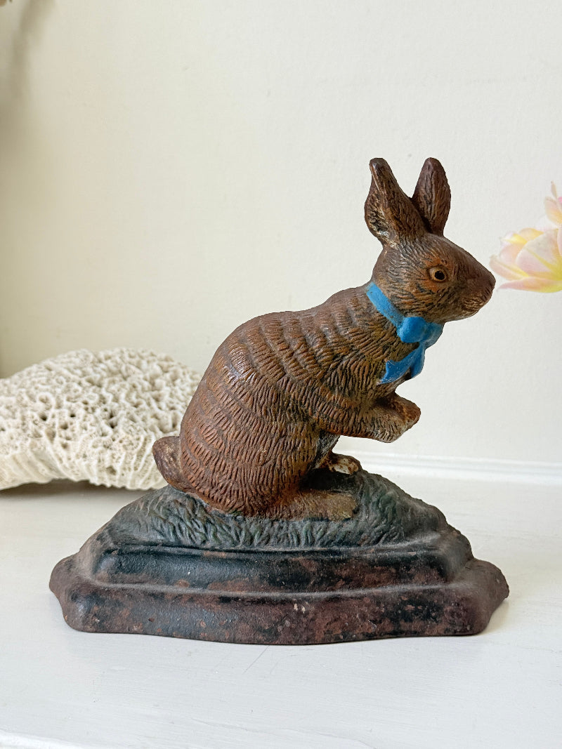 A vintage-style cast iron rabbit figurine wearing a blue bow tie, sitting on a dark textured base against a light beige background.