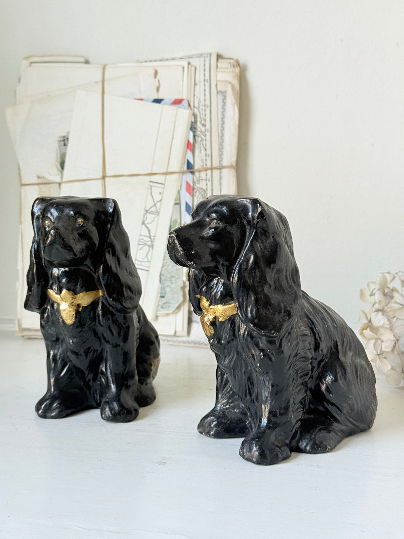 Two glossy black ceramic Cocker Spaniel dog figurines with gold bowties, displayed on a white surface with vintage envelopes in the background.
