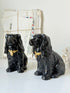 Two glossy black ceramic Cocker Spaniel dog figurines with gold bowties, displayed on a white surface with vintage envelopes in the background.
