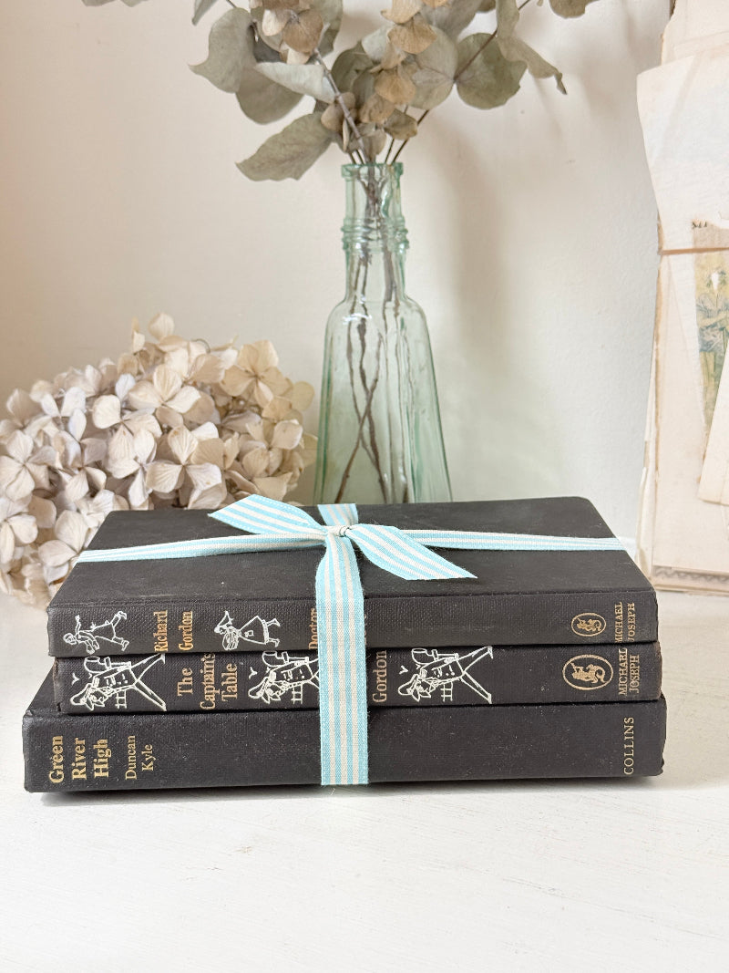 Three antique black books with decorative spines tied with a striped cotton ribbon on a white surface, styled with glass bottles