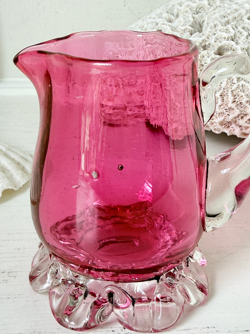 Antique cranberry glass cream jug with clear applied handle, crimped pedestal foot and deep pink hand blown glass body
