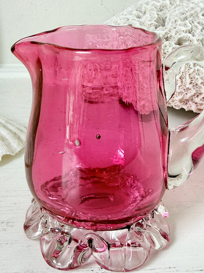 Antique cranberry glass cream jug with clear applied handle, crimped pedestal foot and deep pink hand blown glass body
