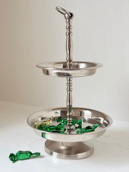 Two tier silver tone cake stand with polished reflective finish styled with green wrapped sweets on a white surface