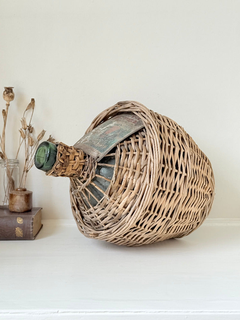 antique French demi john olive oil jar in wicker basket with green glass bottle and painted metal label, displayed on white surface