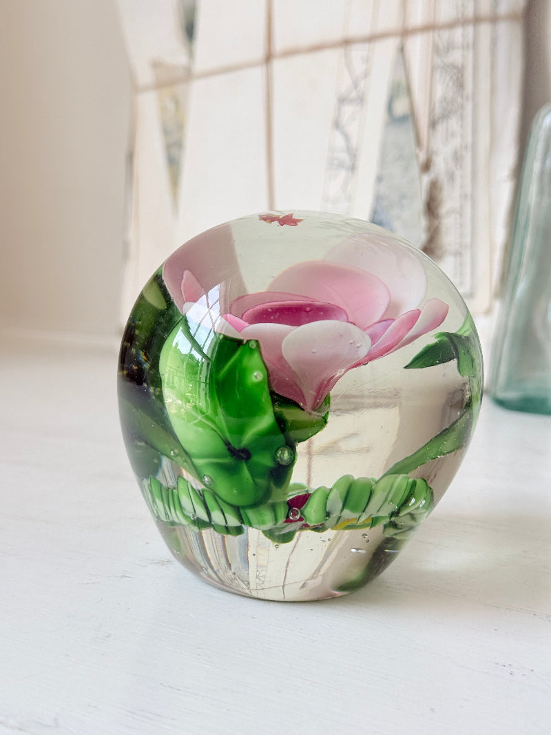 Large clear glass paperweight with a pink flower and green leaves inside, photographed on a white surface