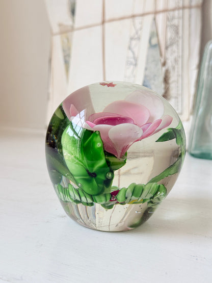 Large clear glass paperweight with a pink flower and green leaves inside, photographed on a white surface