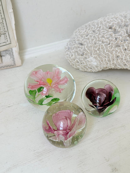 Trio of vintage glass paperweights with hand worked pink and plum floral detail, clear glass domes and green leaf decoration