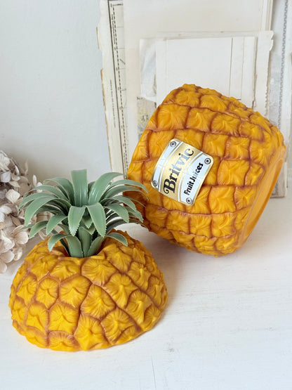 Two decorative pineapple-shaped containers or planters on a white surface, with one upright and one lying down. The upright one has a green faux pineapple crown and a visible label that reads &