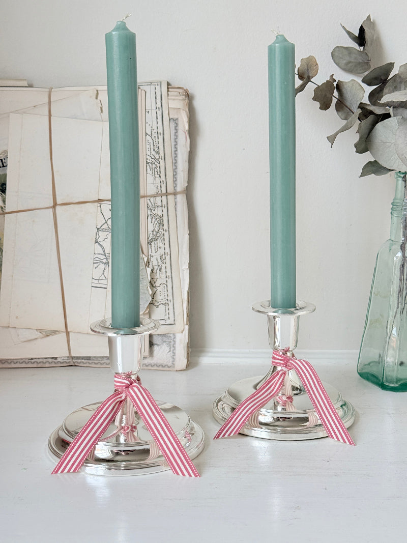 Pair of silver tone candlesticks with wide round stepped bases holding green candles, photographed on a white surface