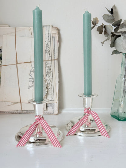 Pair of silver tone candlesticks with wide round stepped bases holding green candles, photographed on a white surface