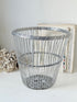 A silver metal wire basket with a woven circular design, styled on a white surface with a vintage frame and dried flowers in the background.