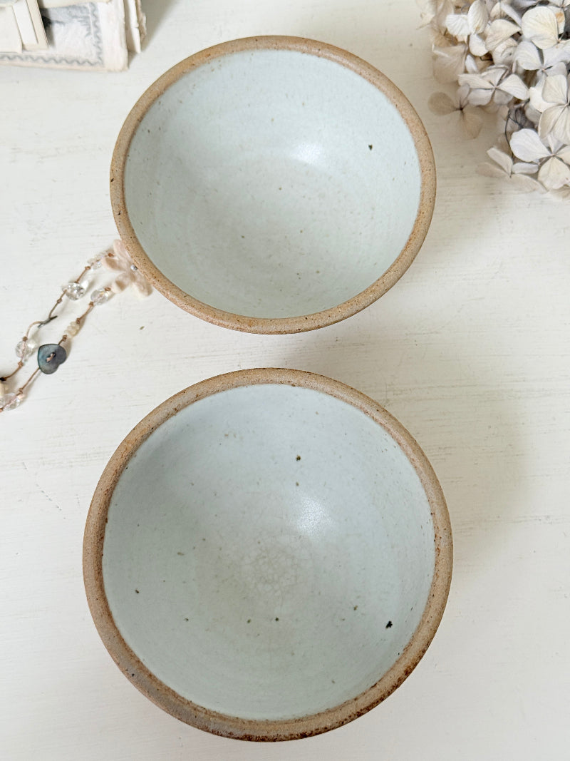 Two handmade ceramic bowls with a light blue-green glazed interior and a rustic brown clay rim, displayed on a white wooden surface with dried flowers and a beaded necklace as props.