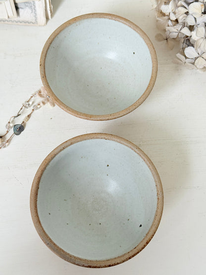 Two handmade ceramic bowls with a light blue-green glazed interior and a rustic brown clay rim, displayed on a white wooden surface with dried flowers and a beaded necklace as props.