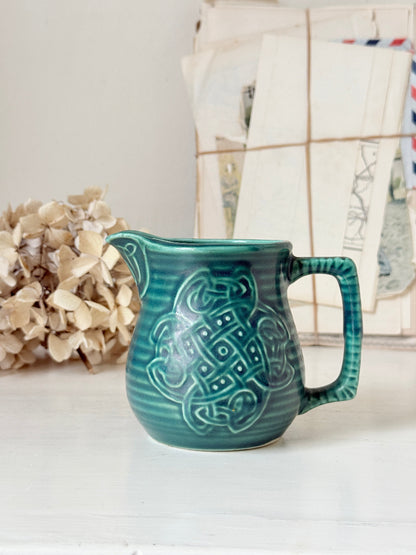 A teal green ceramic pitcher with a raised Celtic knot pattern on the front, set on a white surface with a blurred background of dried flowers and vintage papers.