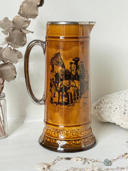 Antique Ridgways stoneware tall jug photographed on a white surface in amber treacle glaze with black transfer scenes and silver tone rim