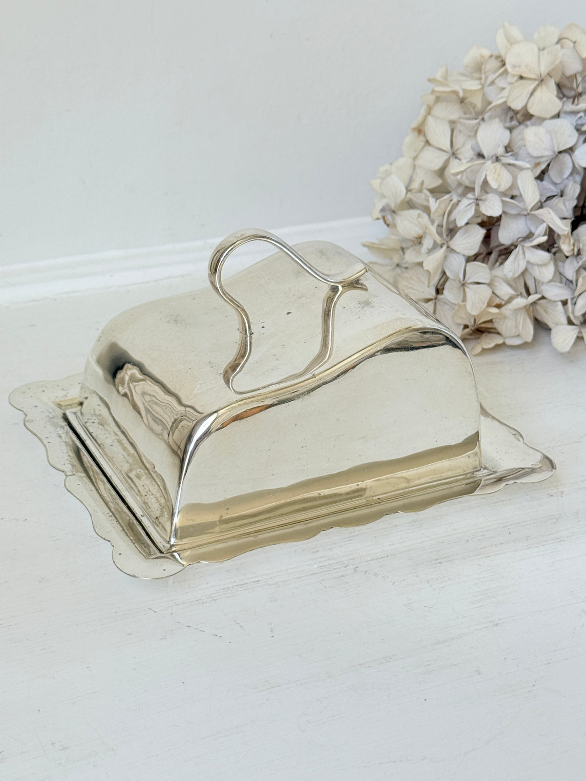 A polished silver butter dish with a hinged lid and scalloped base, shown on a white surface with dried flowers in the background.