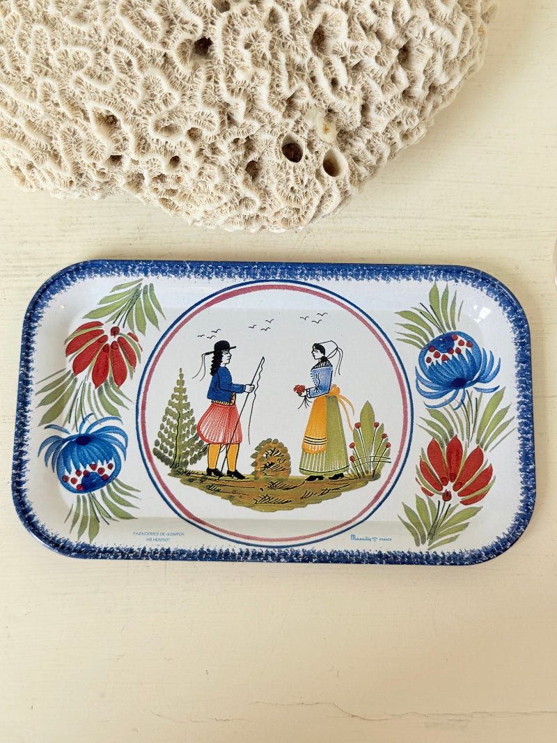 A rectangular enamel tray with rounded corners, featuring a colorful hand-painted folkloric illustration of two figures in a circle, surrounded by floral motifs and a blue patterned border. The tray is displayed on a light beige wooden surface with a textured coral fragment in the background.