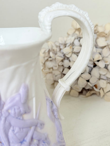 A close-up of a white ceramic pitcher with a decorative handle and light purple embossed figures on the body, set against a soft beige background with dried flowers.