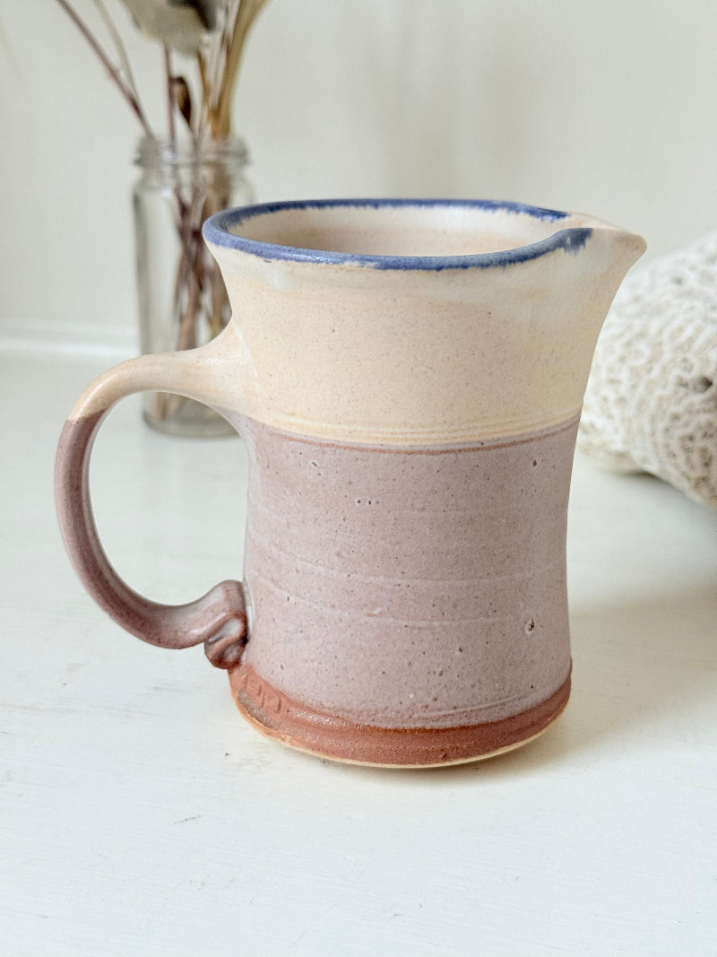 Studio pottery milk jug in cream and lavender glaze with impressed initials, photographed on a white surface.