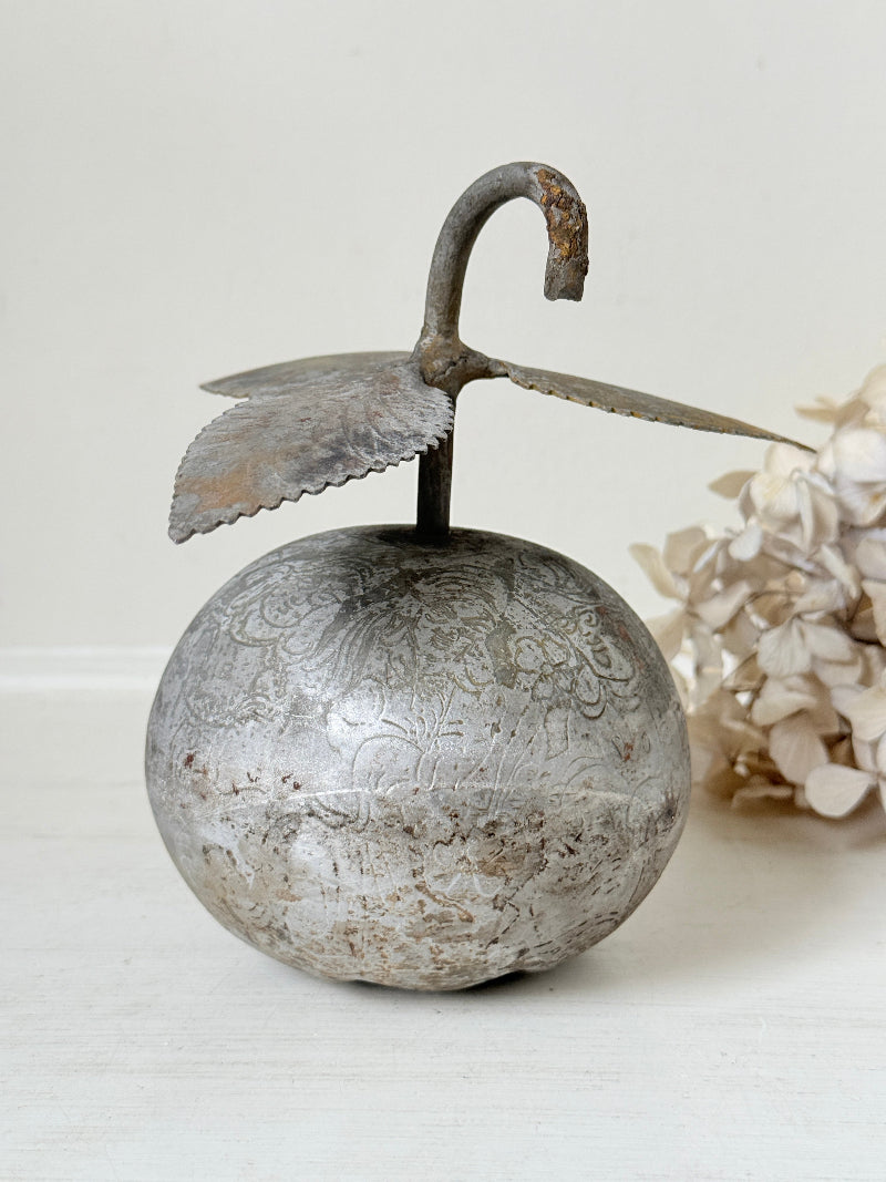 An antique-style metal decorative object shaped like a round silver-toned fruit or ornament with a curved hook handle and leaf-like wings, shown on a light neutral surface with dried flowers in the background.