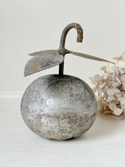 An antique-style metal decorative object shaped like a round silver-toned fruit or ornament with a curved hook handle and leaf-like wings, shown on a light neutral surface with dried flowers in the background.