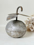 An antique-style metal decorative object shaped like a round silver-toned fruit or ornament with a curved hook handle and leaf-like wings, shown on a light neutral surface with dried flowers in the background.