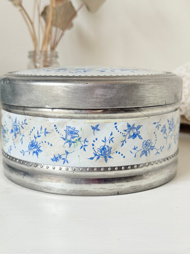 Vintage round shortbread tin with blue floral pattern on silver background, photographed on a white surface.