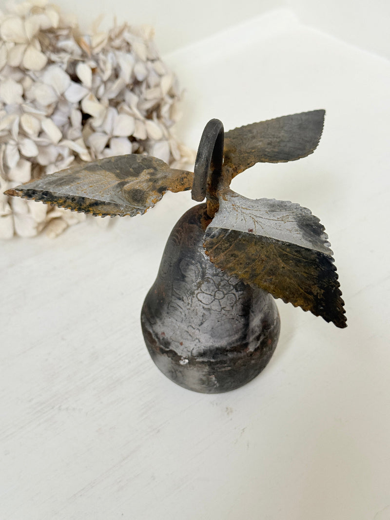 A rustic metal bird bell decoration with a distressed dark gray and brown patina, displayed on a white surface next to dried flowers.